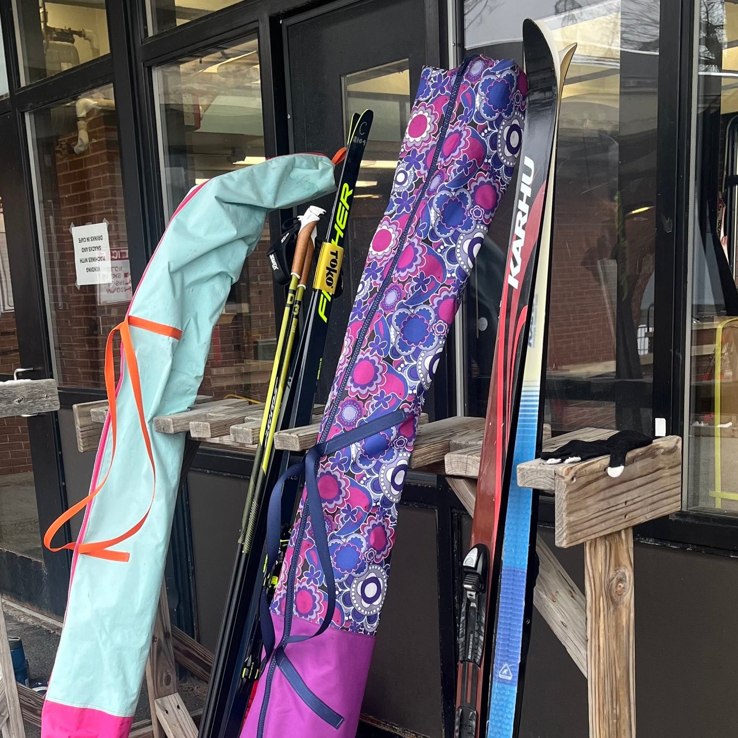 Skis and ski poles with colorful covers leaning against a wooden stand outside a building.
